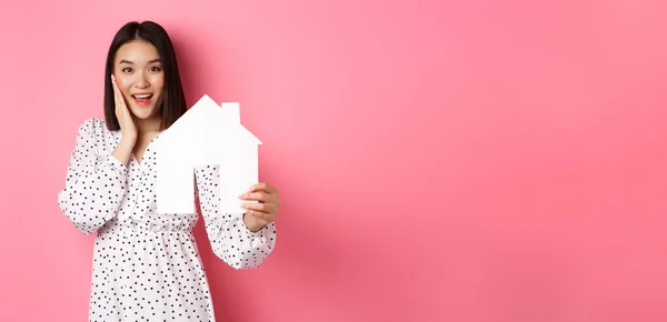 Real estate. Adult asian woman searching for home, holding house model and smiling, promo of broker company, standing over pink background.