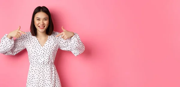 Beautiful korean woman pointing fingers at your logo and smiling, standing in dress over romantic pink background. Copy space