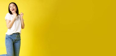 Mutlu Asyalı kadının dans edip mutluluktan atladığı, zaferi kazandığı ve kutladığı, sarı arka planda poz verdiği tam boy bir fotoğraf..