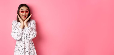 Turizm ve yaşam tarzı konsepti. Sevimli güneş gözlüklü ve elbiseli heyecanlı Asyalı kadın, mutluluk çığlığı atıyor ve kameraya bakıyor, pembe arka planda şaşkın şaşkın duruyor..