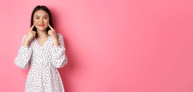 Tatlı, utangaç Asyalı kız yanaklarını dürtüyor, kameraya mutlu bir şekilde gülümsüyor, romantik beyaz elbiseler giyiyor, pembe arka planda duruyor..