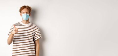 Covid-19, salgın hastalık ve sosyalleşme konsepti. Kızıl saçlı genç adam Coronavirus 'u önlemek için tıbbi maske takıyor. Baş parmak gösteriyor, beyaz arka plan..