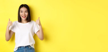 Beyaz tişörtlü, güzel Asyalı bir kadın, baş parmağını kaldırıyor ve gülümsüyor, iyi bir teklif sunuyor, ürün öneriyor, sarı arka planda tatmin olmuş bir şekilde duruyor..