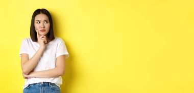 Güzellik ve moda konsepti. Düşünceli Asyalı kadın bir şeyi düşünürken düşünceli görünüyor, sarı arka planda duruyor..