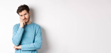 Kuşkucu genç adam sıkılmış yüzüyle seni dinliyor, kameraya gönülsüz bakıyor, beyaz arka planda eşofman üstü duruyor..