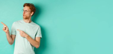 Gözlüklü ve tişörtlü etkileyici kızıl adam, parmaklarını gösterip reklam teklifine bakıyor, mutlu bakıyor, turkuaz arka planda dikiliyor..