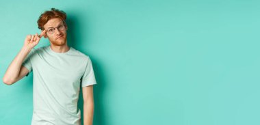 Şüpheci kızıl saçlı bir adam aptal ya da deli birini azarlıyor, parmağıyla kafasını işaret ediyor ve kameraya bakıyor, arka planda duruyor..