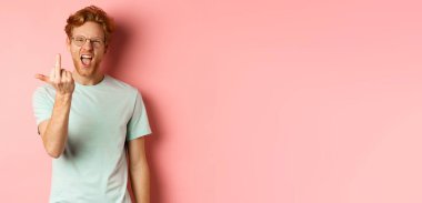 Arrogant and rude redhead man in glasses dont give a fuck, showing middle fingers at camera and frowning, standing over pink background.