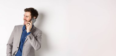 Mutlu iş adamı gülüyor ve gülümsüyor, cep telefonuyla konuşuyor, el ele tutuşuyor ve neşeli yüzüyle boş alana bakıyor, beyaz arka plan.