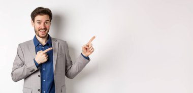 Takım elbiseli mutlu işadamı parmaklarını sağa ve gülümsüyor. Heyecanlı, özel bir anlaşma yapıyor, beyaz arka planda duruyor..