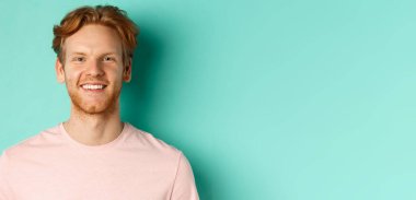Kırmızı sakallı, pembe tişörtlü, beyaz, mükemmel dişleriyle gülümseyen ve turkuaz arka planda duran kızıl saçlı adamı yakından çek..