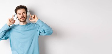 Rahatlamış genç adam favori şarkının tadını çıkarıyor, kulaklıkla müzik dinliyor, gözleri kapalı ve sakin bir yüz, beyaz arka planda duruyor..