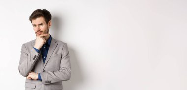 Tereddüt eden takım elbiseli genç işadamı kameraya şüpheci bakıyor, düşünüyor ve karar veriyor, beyaz arka plana karşı duruyor..