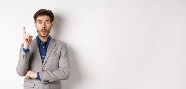 Heyecanlı takım elbiseli işadamı iyi fikir sunuyor, Eureka işaretinde parmağını kaldırıyor ve komik görünüyor, öneriler söylüyor, beyaz arka planda duruyor..