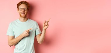 Gözlüklü arkadaş canlısı kızıl saçlı adam sağ üst köşeyi işaret ediyor, reklam afişini gösteriyor ve kameraya gülümsüyor, pembe arka planda duruyor..