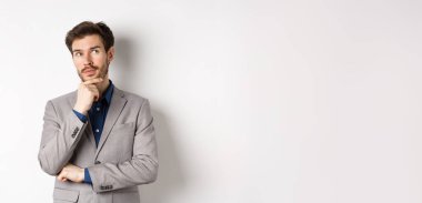 Takım elbiseli dalgın yakışıklı adam yukarı bakıyor ve düşünüyor, seçim yapıyor, fikirleri düşünüyor, beyaz arka plana karşı duruyor..