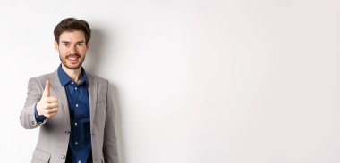 Takım elbiseli yakışıklı iş adamının resmi. Baş parmağını kaldırıyor ve gülümsüyor. Beyaz arka planda duruyor..