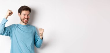 Mutlu sakallı adam zaferi kutluyor, elini kaldırıyor ve zafer kazanıyor, ödül kazanıyor ve sevinçle gülümsüyor, beyaz arka planda duruyor..