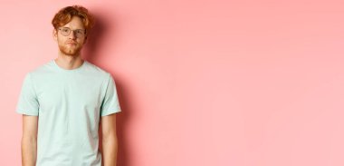 Üzgün ve kasvetli kızıl saçlı, tişörtlü ve gözlüklü adam, kameraya bakıyor sıkılmış ve eğlenmemiş, pembe arka planda duruyor..