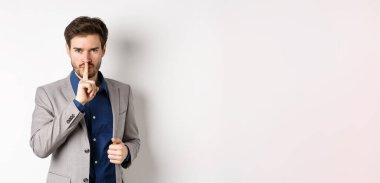 Kızgın işadamı tabu işareti gösteriyor, sessiz olmasını söylüyor, sessizliğe ihtiyacı var, parmaklarını dudaklarına dayayıp kameraya bakıyor, beyaz arka plan.