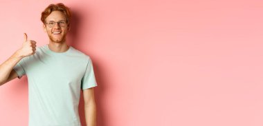Gözlüklü ve tişörtlü neşeli kızıl adam, tatmin olmuş yüzüyle başparmağını kaldırıyor, olumlu tepki gösteriyor, onaylıyor ve seninle aynı fikirde, pembe arka planda duruyor..