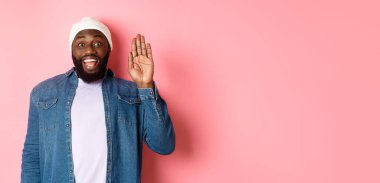 Dostça gülümseyen siyah adam merhaba diyor, el sallıyor, seni selamlıyor, pembe arka planda duruyor..