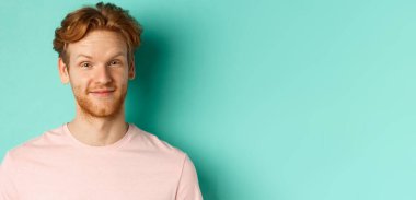 Kızıl sakallı adam memnun görünüyor, onaylıyor ve gülümsüyor, pembe tişörtle turkuaz arka planda duruyor..