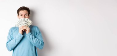 Şaşırmış ve şaşırmış yakışıklı adam para gösteriyor, tanıtım teklifine bakıyor, nakit parayla alışverişe gidiyor, beyaz arka planda duruyor..