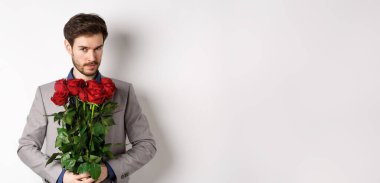 Yakışıklı, maço bir adam, takım elbiseli, elinde bir buket kırmızı gülle kameraya gülümsüyor, sevgililer günü hediyesi hazırlıyor, beyaz arka plan..