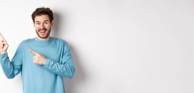 Mavi kazaklı mutlu bir beyaz adam, sol üst köşeyi işaret ediyor, beyaz arka planda bağlantı ya da logo gösteriyor, kameraya gülümsüyor..