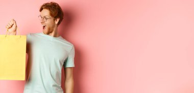 Yakışıklı genç adam hediyeler alıyor, alışveriş çantası tutuyor ve eğleniyor görünüyor, pembe arka planda duruyor..