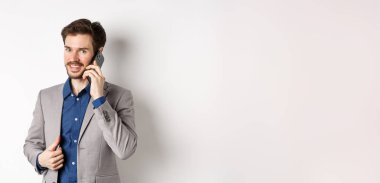 Başarılı iş adamı takım elbisesini onarıyor ve cep telefonuyla konuşuyor. Kameraya gülümsüyor, beyaz arka planda duruyor..