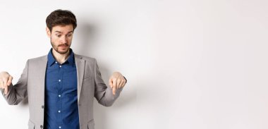 Şaşkın iş adamı yukarı kaş kaldırarak aşağı bakıyor ve tuhaf bir şey görüyor, takım elbiseli beyaz arka planda duruyor..