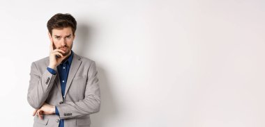 Sıkıntılı, kaşları çatık, takım elbiseli bir iş adamı kameraya bakıp düşünüyor ya da karar veriyor, beyaz arka planda duruyor..