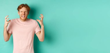 Annoyed and pissed-off redhead guy looking angry, shouting and grimacing, shaking hands aggressive, standing furious against mint background.