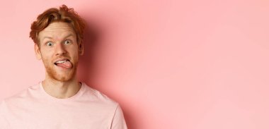 Komik kızıl saçlı adamın dil göstermesi, kameraya aptal suratlar yapması, pembe arka planda neşeli bir şekilde dikilmesi..