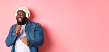 Mutlu Afro-Amerikalı adam dans ediyor, kulaklıkla müzik dinliyor ve pembe arka planda kameraya bakıyor..