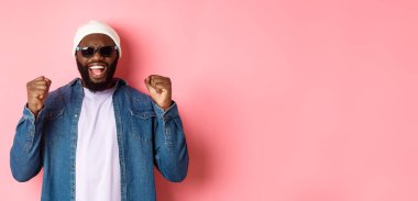 Güneş gözlüklü tatmin olmuş Afro-Amerikalı adam evet diyor, sevinçle yumruk tokuşturuyor, başarıyı kutluyor, pembe arka planda kazanan gibi duruyor..