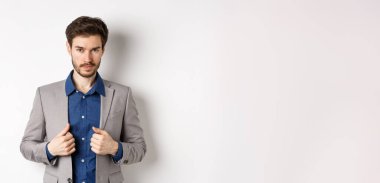 Sakal düzeltme kıyafeti giymiş, kameraya küstah bakışlı, hazır ve kararlı, beyaz arka planda duran kendine güvenen genç bir adam..