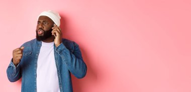 Kızgın Afro-Amerikalı adam telefonda konuşuyor ve birini yumruğuyla tehdit ediyor, rahatsız bir şekilde bakıyor, pembe arka planda duruyor..