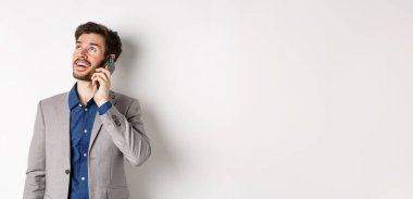 Tasasız genç adam cep telefonuyla konuşuyor ve gülümsüyor, rüya gibi bir yüzle yukarı bakıyor, beyaz arka planda takım elbise içinde duruyor..