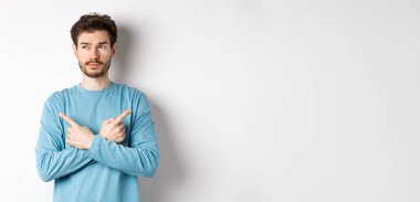 Kararsız adam seçim yapıyor, parmaklarını iki varyanta doğrultuyor ve karar veriyor, sol tarafta dalgın dalgın bakıyor, beyaz arka planda duruyor..