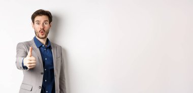 Takım elbiseli yakışıklı iş adamının görüntüsü. Heyecanlı yüzüyle baş parmağını gösteriyor. Beyaz arka planda duruyor..