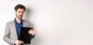 Genç erkek CEO elinde belgelerle pano tutuyor ve kameraya bakıyor, ofisi inceliyor, beyaz arka planda duruyor..