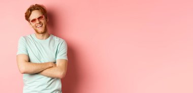 Turizm ve tatil konsepti. Kızıl saçlı, güneş gözlüklü ve tişörtlü, göğsünde çapraz kolları olan ve kameraya neşeyle gülümseyen, pembe arka planlı arkadaş canlısı bir genç..