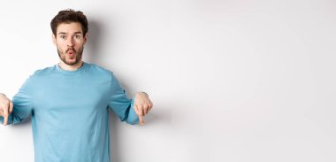 Sakallı, sıradan mavi tişörtlü, hayret verici ve aşağıdan işaret eden genç adam, şuraya bak, beyaz arka planda duruyor..