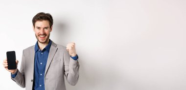 E-ticaret ve online alışveriş konsepti. Adam internette para kazanıyor, akıllı telefon ekranı ve kazanan hareketi gösteriyor, gülümsüyor, beyaz arka planda duruyor..