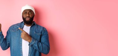 Şüpheci ve şüpheci siyah adam kameraya bakıyor emin değil, parmaklarını sol üst köşedeki belirsiz bir şeye doğrultuyor, pembe arka planda duruyor..