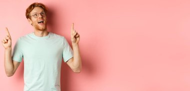 Harika gözlüklü kızıl saçlı adam özel indirimlere bakıyor ve parmaklarını şaşkın şaşkın yukarı kaldırıyor. Pembe arka planda duruyor..