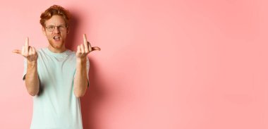 Arrogant and rude redhead man in glasses dont give a fuck, showing middle fingers at camera and frowning, standing over pink background.